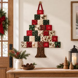 Puede incluir: Una decoración de pared con forma de árbol de Navidad hecha de cuadrados de tela acolchada en colores rojo, verde y crema. Los cuadrados presentan patrones de flores de pascua, acebo, copos de nieve y un ciervo. Un tronco de tela marrón está unido a la base.