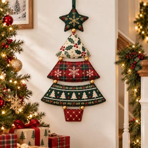 Op de afbeelding: Een wanddecoratie in de vorm van een kerstboom, bestaande uit vijf gestapelde, gewatteerde stoffen segmenten in donkergroen, rood en crème. De delen zijn voorzien van geborduurde sneeuwvlokken, hulstbladeren en kleine boommotieven, verbonden door goudkleurige bollen.