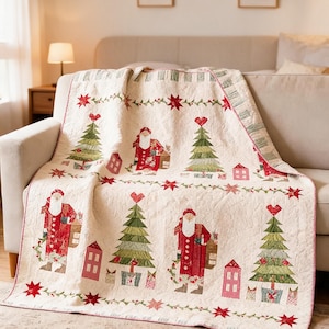 May include: A cream-coloured quilted throw blanket featuring a repeating pattern of Santa Claus figures, green Christmas trees topped with red hearts, and small pink houses. The design includes red star accents and a decorative border of green vines.