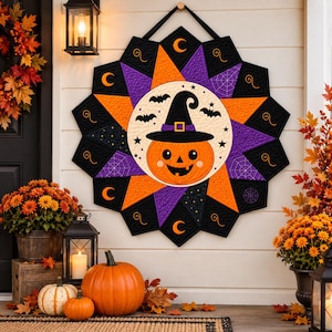 Puede incluir: Un tapiz acolchado con un panel circular central que muestra una calabaza sonriente con sombrero de bruja, rodeado por un borde en forma de estrella en negro, naranja y morado con patrones de lunas crecientes, telarañas y murciélagos.