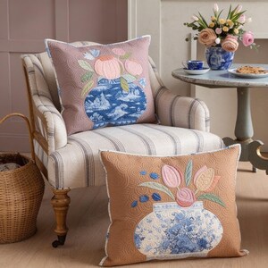 Könnte beinhalten: Zwei quadratische Zierkissen mit gesteppten Oberflächen und Stickereien von blau-weißen Vasen, gefüllt mit rosa und pfirsichfarbenen Blumen. Ein Kissen ist malvenfarben, das andere hellbraun, beide mit detaillierten, geschwungenen Steppmustern.