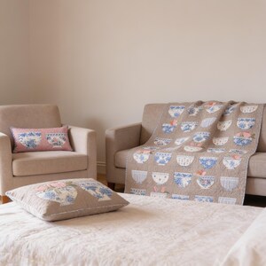 May include: A beige quilt and two matching throw pillows featuring a repeating pattern of blue and white bowls with floral accents. One pillow is rectangular with a pink background, while the other is square with a beige background.