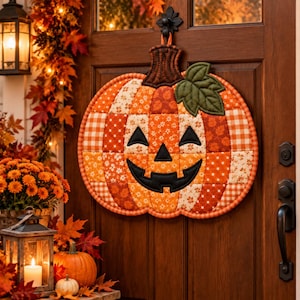 May include: A quilted fabric pumpkin wall hanging featuring a patchwork design of orange, brown, and cream fabrics with various patterns. The pumpkin has a black stitched jack-o'-lantern face, a brown stem, and a single green fabric leaf attached.