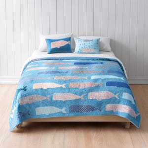 Peut inclure: Une courtepointe bleue ornée d'un motif répétitif de baleines dans diverses nuances de bleu, de blanc et d'orange avec des textures et des imprimés variés. Deux oreillers rectangulaires assortis sont placés en tête de lit avec des motifs.
