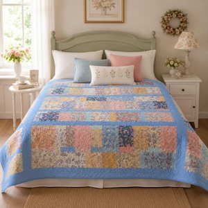 May include: A patchwork quilt featuring a repeating pattern of floral fabric rectangles in shades of blue, pink, and yellow, bordered by a solid light blue fabric. The quilt is displayed on a bed with white and pink pillows and a blue accent pillow.