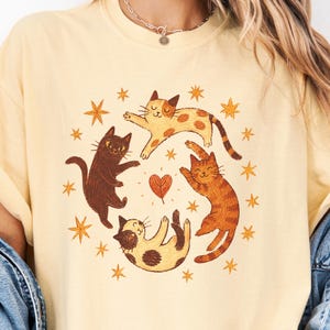 May include: A pale yellow t-shirt featuring a circular graphic of four cats in various colors and patterns, including black, orange tabby, and spotted, surrounding a central heart shape, all accented by small orange star illustrations.