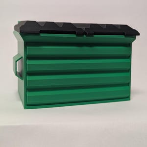 May include: A small green dumpster model with a black hinged lid. The container features a rectangular body with horizontal ribbed panels and a handle on the side. The design mimics a standard industrial waste bin with a two-part lid structure.
