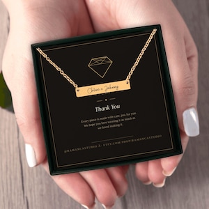 Puede incluir: Un collar de barra en tono dorado con el texto grabado Chloe x Johnny, presentado en una caja de regalo negra. El inserto de la caja muestra un icono de diamante, el texto Thank You e información de la marca, incluyendo una URL y un usuario.