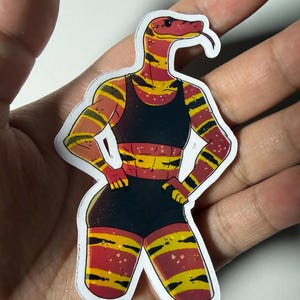 Peut inclure: Un autocollant découpé représentant un personnage serpent anthropomorphe à la peau rayée rouge et jaune, portant une brassière de sport noire et un short de sport noir. Le personnage a une musculature athlétique, une tête de serpent et les mains sur les hanches.