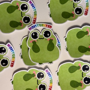Peut inclure: Une collection d'autocollants découpés représentant une grenouille de dessin animé avec de grands yeux noirs brillants, des joues roses et une peau verte. Chaque autocollant porte le texte MOISTURIZED en dégradé arc-en-ciel au-dessus.