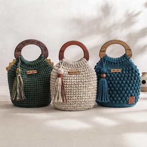 May include: Three handmade crochet bags with round wooden handles. The left bag is dark green with a multi-colored tassel. The center bag is beige with a speckled texture and a tassel. The right bag is teal with a textured pattern and a teal tassel.