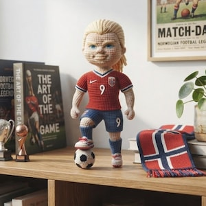 Puede incluir: Una muñeca de ganchillo con cabello rubio que viste una camiseta roja con el número 9 y un logotipo blanco, pantalones cortos azules con el número 9 y calcetines azules. La muñeca está sobre un balón de fútbol blanco y negro. Hay una bufanda cerca.