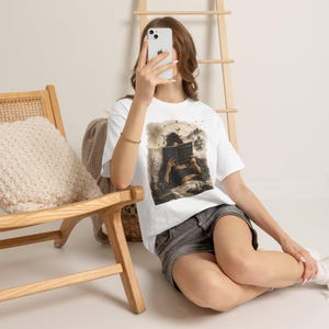 May include: A white short-sleeve t-shirt featuring a graphic print of a person reading a book amidst a mystical landscape with celestial elements, paired with gray denim shorts. The design includes dark, muted tones and intricate line art details.