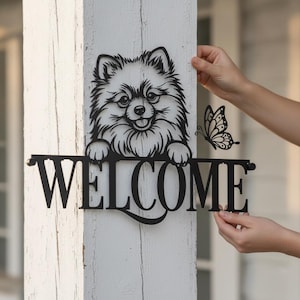 Pode incluir: Uma placa de parede em metal preto com uma ilustração estilizada de um cão Lulu da Pomerânia fofo espreitando sobre a palavra WELCOME em letras maiúsculas em negrito, com um pequeno detalhe de borboleta à direita da cabeça.
