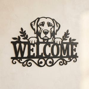 Könnte beinhalten: Ein schwarzes Metall-Wandschild mit dem Kopf eines Labrador-Hundes, dessen Pfoten auf dem Wort WELCOME in Großbuchstaben ruhen, akzentuiert durch blättrige Zweige an beiden Seiten und dekorative Schnörkel entlang der unteren Kante des Schildes.