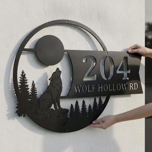 Könnte beinhalten: Ein rundes Adressschild aus schwarzem Metall mit einem Scherenschnitt-Design eines heulenden Wolfs auf einem Felsen, Tannen und einem Vollmond. Das Schild zeigt die Nummer 204 und den Text WOLF HOLLOW RD in einer fetten, serifenlosen Schrift.
