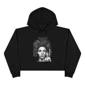 May include: A black hooded sweatshirt featuring a graphic print on the chest. The design includes the text "SHE IS:" and "FEARLESS" alongside a detailed illustration of a person with intricate hair patterns and a smaller figure beside them.