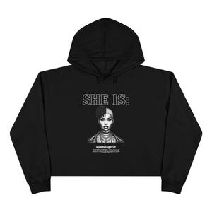 May include: A black cropped hoodie featuring a white graphic print on the chest. The design includes the text 'SHE IS:' above a black and white illustration of a person with a split hairstyle, and the word 'Unapologetic' with smaller text below.