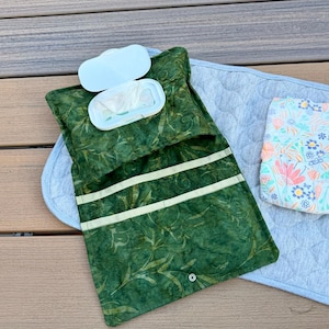 May include: A green fabric diaper clutch with a batik-style leaf pattern, featuring an integrated white plastic wipes case, two internal pockets, and a metal snap closure, accompanied by a folded floral patterned diaper on a gray quilted mat.