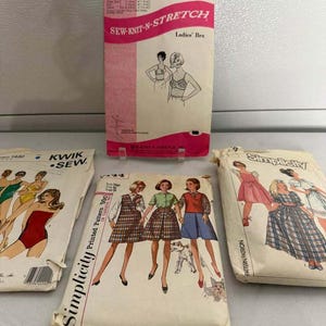 May include: Four vintage sewing pattern envelopes featuring illustrations of clothing. One envelope is for a Sew-Knit-N-Stretch bra, two are Simplicity patterns showing dresses and skirts, and one is a Kwik Sew pattern displaying various swimsuits.