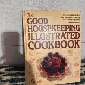 May include: The Good Housekeeping Illustrated Cookbook with a cream-coloured cover and dark brown text. The title is printed in large letters, and the cover features a photograph of a cream puff ring topped with strawberries and chocolate glaze.