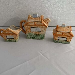 May include: A ceramic tea set consisting of a teapot, a creamer, and a sugar bowl, all shaped like small cottages with thatched roofs, tan walls, green shrubbery at the base, and rectangular windows with black frames and white panes.