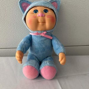 May include: A plush doll with a light blue cat-themed hood featuring pink inner ears. The doll has blue eyes, a pink nose, and rosy cheeks. It wears a matching light blue fuzzy onesie with a pink ribbon tied at the neck and pink fabric feet.