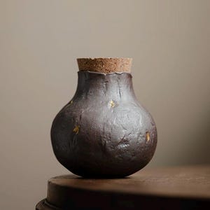 May include: A small, bulbous, dark brown ceramic vessel with a textured, uneven surface and scattered gold-colored flecks. The container features a narrow neck topped with a cylindrical, light brown cork stopper that fits snugly into the opening.