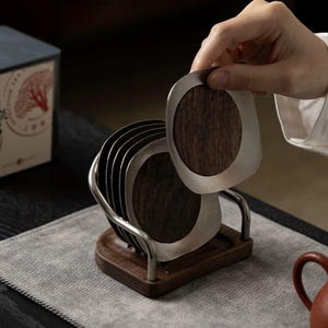 May include: A set of circular coasters featuring dark brown wood centres surrounded by brushed silver-toned metal frames. The coasters are stored in a rectangular wooden base with a curved metal rack that holds them in an upright vertical position.