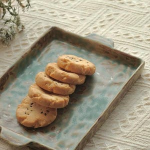Puede incluir: Una bandeja rectangular de cerámica con un esmalte moteado en azul y marrón, que cuenta con dos pequeñas asas en los lados. Cinco galletas redondas de color claro con motas oscuras visibles están dispuestas en una fila superpuesta.
