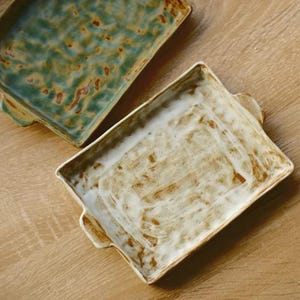 May include: Two rectangular ceramic baking dishes with side handles. One dish features a mottled teal and brown glaze with a textured surface, while the other displays a creamy white glaze with visible brown brushstrokes and a smooth finish.