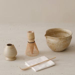 May include: A matcha tea set consisting of a beige ceramic whisk holder, a bamboo whisk with a black thread, a beige ceramic pouring bowl with a textured finish, a bamboo tea scoop, and a folded white cloth napkin arranged on a flat surface.