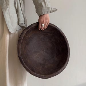May include: A large, circular, dark brown wooden bowl with a visible grain pattern and a smooth, rounded rim. The bowl features a deep, concave interior and a weathered, matte finish, showcasing the natural texture and dark tones of the wood.