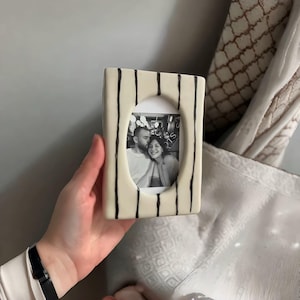 Puede incluir: Un marco de fotos rectangular de cerámica con una base de color blanco roto y rayas verticales negras pintadas a mano. El marco presenta una abertura ovalada central que contiene una fotografía en blanco y negro de dos personas sonriendo.
