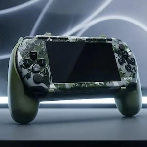 以下が含まれることがあります： 緑、黒、白の迷彩柄が施された携帯型ゲーム機。中央に画面があり、黒い操作ボタンと側面に無地のオリーブグリーンのグリップを備えています。前面にはSonyのロゴが表示されています。