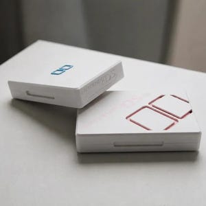Puede incluir: Dos consolas de videojuegos portátiles Nintendo DS blancas apiladas una sobre otra. La consola superior presenta un logotipo DS azul, mientras que la inferior muestra un logotipo DS rosa. Ambos dispositivos tienen forma rectangular.