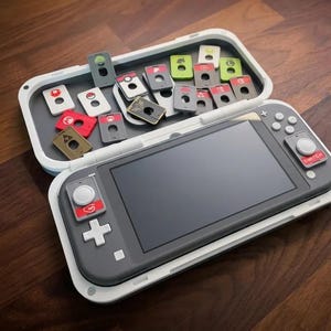 May include: A gray handheld gaming console with a black screen, white buttons, and a red Switch label, resting in a white open carrying case. The case interior contains multiple small, rectangular game cartridges featuring various colorful icons.