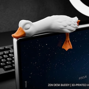 Könnte beinhalten: Eine weiße, schlafende Gänsefigur mit orangefarbenem Schnabel und orangefarbenen Schwimmfüßen, die auf der Oberkante eines Computermonitors ruht. Der Text auf dem Bildschirm lautet: ZEN DESK BUDDY | 3D-PRINTED ARTISAN | SLEEPING GOOSE | For Laptops & Monitors.