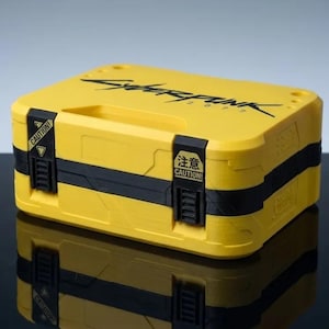 May include: A rectangular yellow storage case with black accents and a handle. The top features the black text Cyberpunk 2077. Two black latches on the front are labeled with yellow caution stickers, one including the Japanese text 注意 and the word CAUTION.