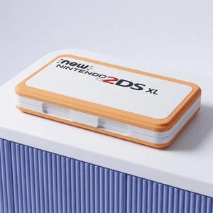 Puede incluir: Una consola de juegos portátil New Nintendo 2DS XL blanca y naranja en posición cerrada. La cubierta superior presenta el texto New Nintendo 2DS XL con un pequeño logotipo multicolor sobre la palabra New. El dispositivo tiene forma rectangular con esquinas redondeadas.