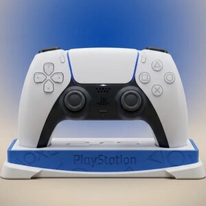 May include: A white and black wireless video game controller sits on a white and blue charging dock. The dock features the word PlayStation embossed on the front, along with the iconic triangle, circle, cross, and square symbols on the base.