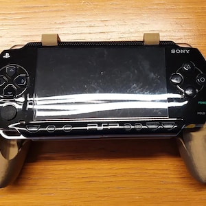 May include: A black Sony PSP handheld gaming console mounted in a bronze-colored plastic ergonomic grip. The device features a central screen, directional pad, action buttons, and the text SONY and PSP printed on the front face of the console.