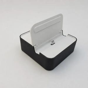 Puede incluir: Una base de carga en blanco y negro con una base rectangular, una superficie superior blanca y un panel de soporte vertical central. Un pequeño conector USB-C metálico sobresale del centro de la base blanca para conectar un dispositivo.