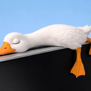 May include: A white duck figurine with a bright orange beak and orange webbed feet, depicted in a sleeping position with its head resting on the top edge of a black computer monitor, while its feet hang down over the side of the screen frame.