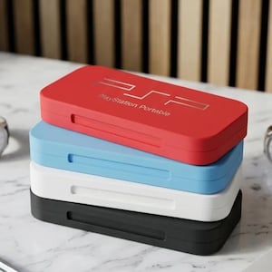 Op de afbeelding: Een stapel van vier rechthoekige PlayStation Portable-hoesjes in rood, lichtblauw, wit en zwart. Het bovenste rode hoesje is voorzien van het zilveren PSP-logo en de tekst PlayStation Portable. Elk hoesje heeft een horizontale uitsparing.