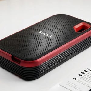 Könnte beinhalten: Ein rechteckiges SanDisk Gehäuse für tragbare SSDs mit einer schwarzen, texturierten Oberfläche und einem perforierten Muster. Das Gehäuse hat einen metallisch roten Rand und ein schwarzes, geripptes Seitendesign mit einer Aussparung.