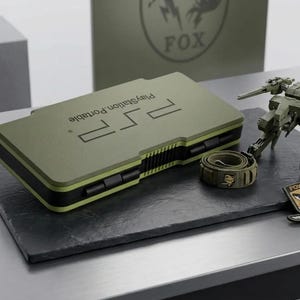 Pode incluir: Um estojo de console PlayStation Portable na cor verde-oliva com detalhes em preto, contendo o texto PlayStation Portable. Ao lado, há uma pequena figura robótica verde-oliva, uma alça enrolada com um logotipo e um emblema Foxhound.