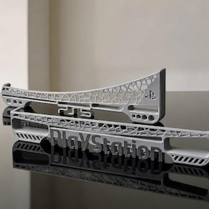 May include: Two light gray 3D-printed console accessories featuring a complex lattice structure. One piece displays the PS5 logo, while the other features the PlayStation wordmark. Both items have a matte finish and geometric cutouts.