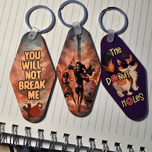 Pode incluir: Três porta-chaves em forma de diamante com argolas de metal. O primeiro mostra um céu nublado, uma bomba e o texto YOU WILL NOT BREAK ME. O segundo apresenta uma cena de fantasia com uma figura. O terceiro mostra um gato com óculos de sol e o texto THE DONUT HOLES.