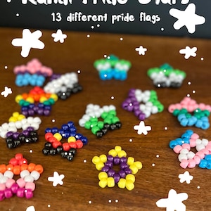 May include: A collection of star-shaped ornaments crafted from colorful plastic pony beads, arranged to represent 13 different pride flags. The text Kandi Pride Stars, 13 different pride flags, and DarkCottageCorner are visible.